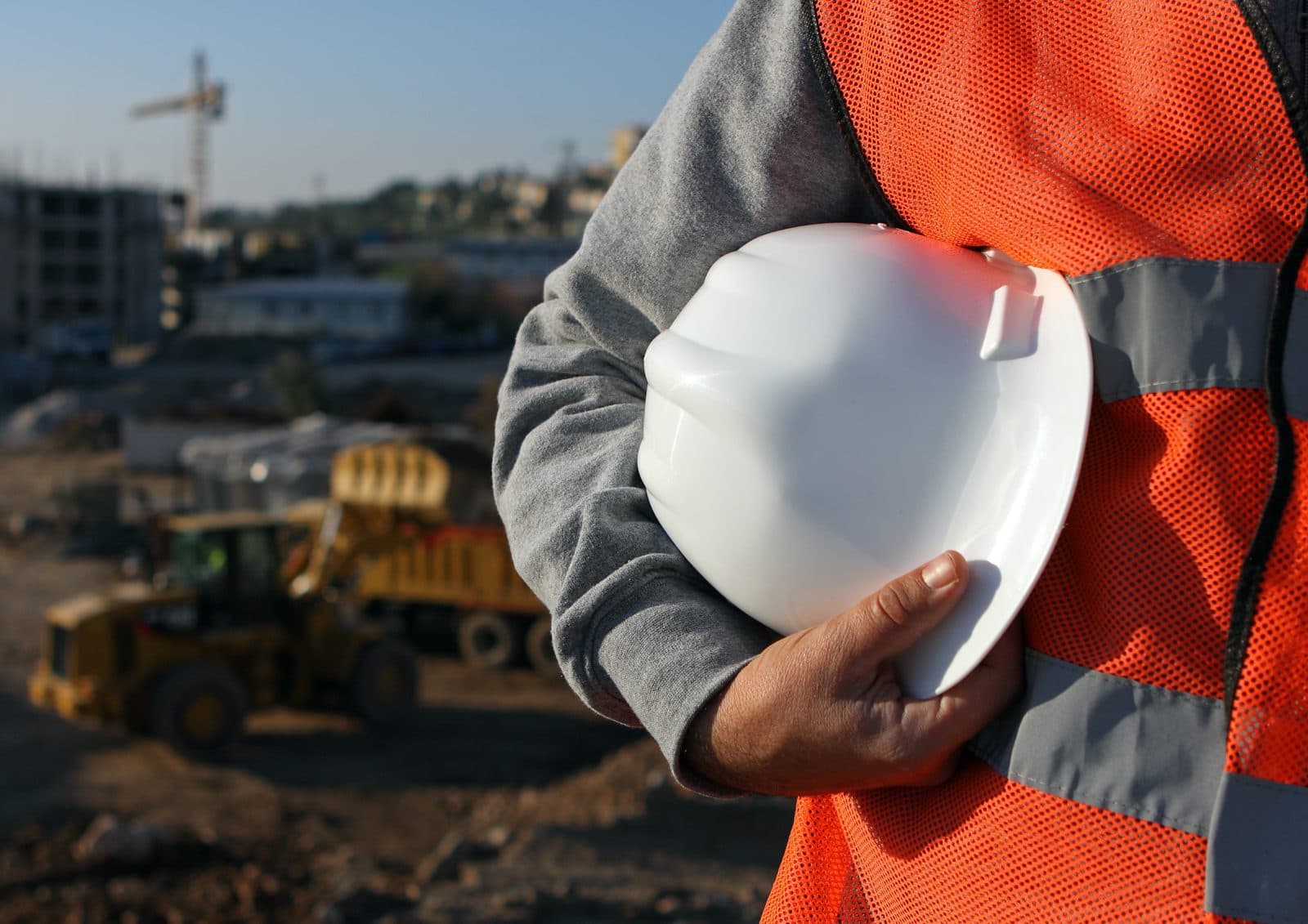 Construction professional in a safety vest holding a white hard hat at an active job site